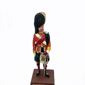 May include: A detailed figurine of a Gordon Highlander soldier, wearing a red jacket, black hat, and kilt. The figurine stands on a brown base with the text "GORDON HIGHLANDERS". The soldier holds a sword and a sporran.