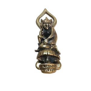Könnte beinhalten: Eine metallene, messingfarbene Figur, die ein sitzendes Wesen mit spitzen Ohren auf einem Pilz zeigt. Die Basis des Objekts trägt den geprägten Text EXMOOR PIXIE. Das Design umfasst ein bogenförmiges Element über dem Kopf des Wesens.