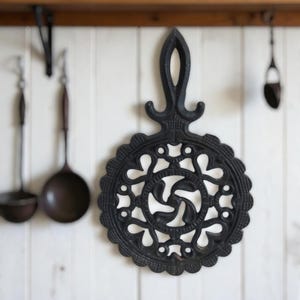 Puede incluir: Salvamanteles de hierro fundido negro con un diseño calado decorativo. El salvamanteles tiene forma circular con asa y bordes festoneados. Utensilios de cocina cuelgan de la pared blanca del fondo.