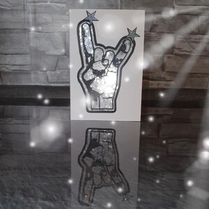 May include: A white card with a silver glitter rock and roll hand gesture with two silver glitter stars on top. The card is reflected in a shiny surface.
