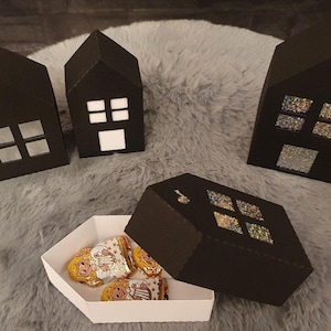 Puede incluir: Cuatro cajas negras de papel con forma de casa con ventanas recortadas, algunas con material reflectante. Una caja está abierta, revelando caramelos envueltos dentro de un recipiente hexagonal blanco. Las casas están sobre una superficie gris y peluda.