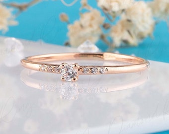 May include: A slender rose gold ring featuring a central round diamond and smaller accent stones. The ring is displayed on a white surface with a blue backdrop and floral details, highlighting its refined aesthetic.