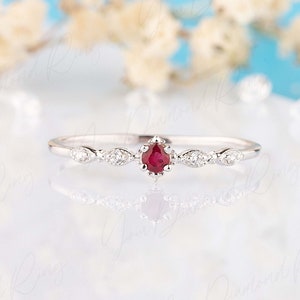 Op de afbeelding: Een delicate zilveren ring met een kleine rode edelsteen omringd door kleine diamanten. De ring ligt op een witte achtergrond.