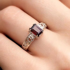 Peut inclure: Une bague dorée ornée d'une pierre rouge foncé de forme rectangulaire. La bague présente un motif floral orné sur le corps. La pierre est sertie dans un cadre carré. La bague est portée au doigt.