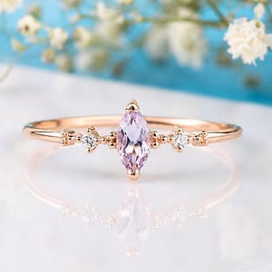 Könnte beinhalten: Ein roségoldener Ring mit einem hellrosa, marquiseförmigen Mittelstein. Das Band ist mit kleinen, runden, klaren Edelsteinen auf beiden Seiten des Mittelsteins verziert. Der Ring liegt auf einer weißen Oberfläche mit einem verschwommenen blau-weißen Blumengrund.