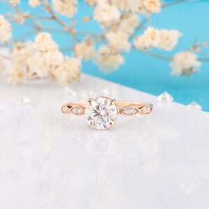 May include: A rose gold ring featuring a large round brilliant-cut center stone held by four prongs. The band is accented with marquise-shaped settings, each containing small round stones, creating a delicate and symmetrical design.