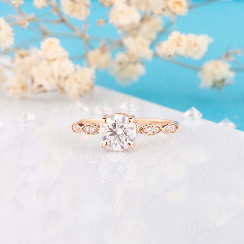 May include: A rose gold ring featuring a large round brilliant-cut center stone held by four prongs. The band is accented with marquise-shaped settings, each containing small round stones, creating a delicate and symmetrical design.