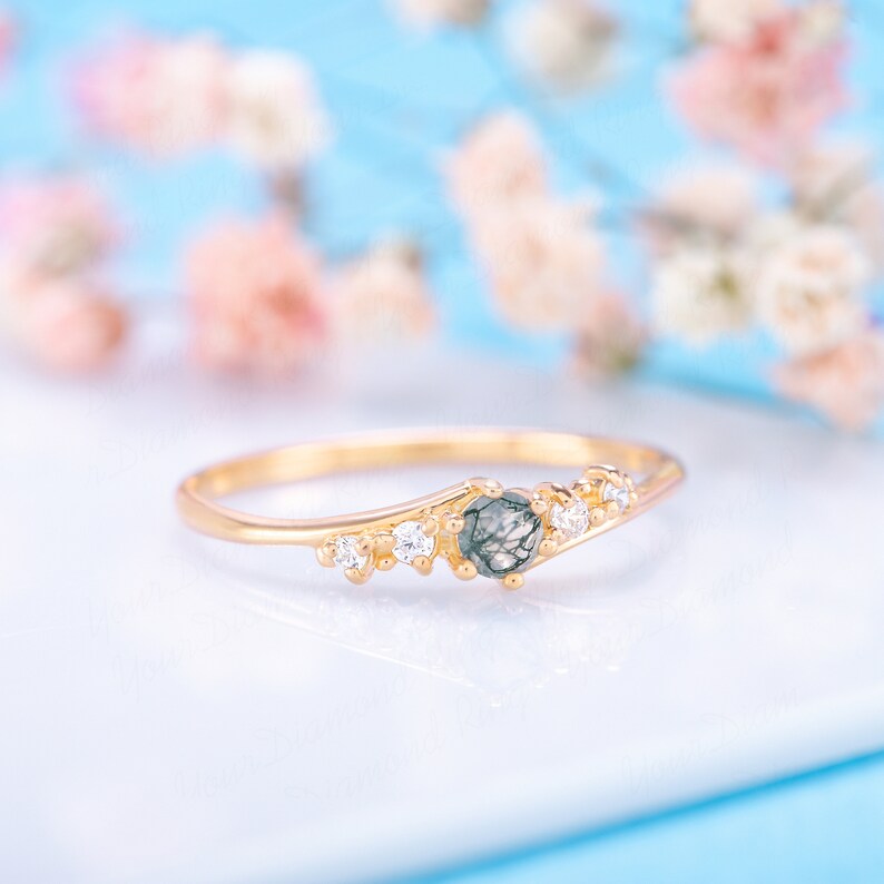 Pode incluir: Um anel de ouro com uma pedra preciosa preta e cinza e pequenas pedras preciosas transparentes. O anel est&aacute; sobre uma superf&iacute;cie branca.