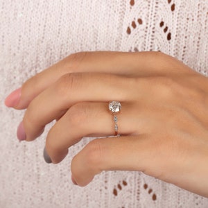 May include: A rose gold ring featuring a large round brilliant-cut center stone held in a four-prong setting. The band is accented with two smaller marquise-cut stones on each side of the center setting, creating a delicate and refined appearance.