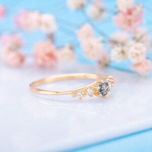 Pode incluir: Um delicado anel de ouro com uma gema verde escura e pequenas pedras transparentes. O anel est&aacute; colocado sobre uma superf&iacute;cie branca com um fundo desfocado de flores cor-de-rosa.