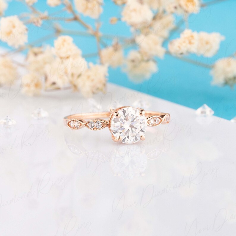 May include: A rose gold ring featuring a large round brilliant-cut center diamond held by four prongs. The band is accented with smaller round diamonds set in marquise-shaped metal frames along the shank, creating a delicate and detailed appearance.