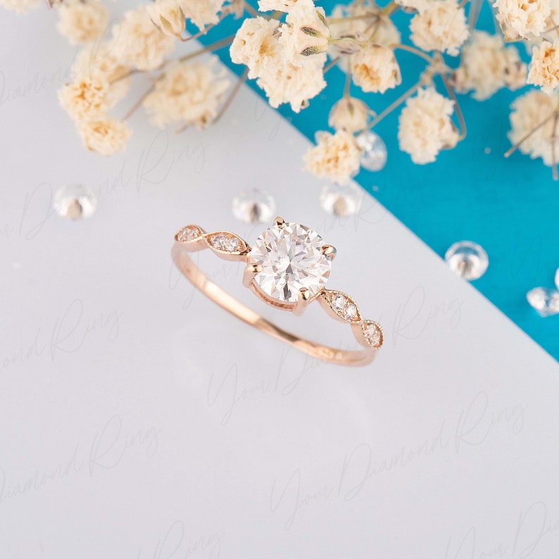 May include: A rose gold engagement ring with a round brilliant cut diamond center stone and a delicate pave diamond band. The ring is set on a white background with white flowers.