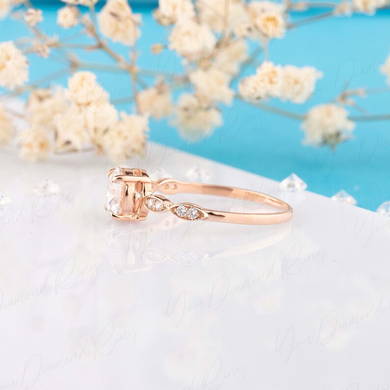 May include: A rose gold ring featuring a central round-cut clear stone held in a four-prong setting. The band displays a delicate, openwork design on the sides, accented with small, round, clear stones embedded along the shank of the ring.
