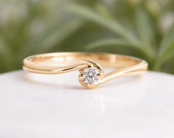 May include: A gold ring with a small, round, clear gemstone at the center. The band curves around the gemstone in a flowing design. The ring is set against a blurred green and white background.