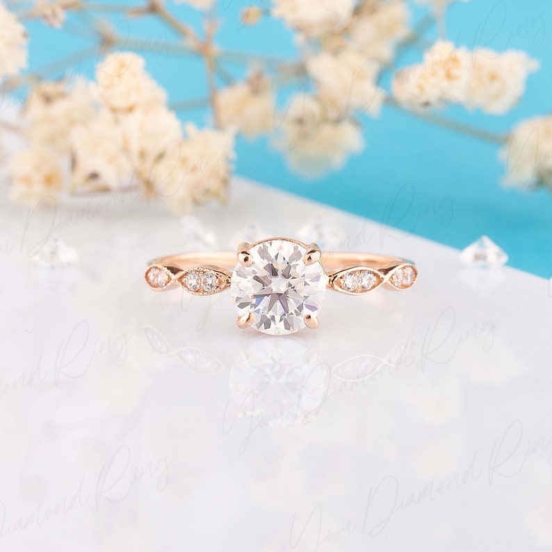 May include: A rose gold ring featuring a large round brilliant-cut center stone held by four prongs. The band is accented with marquise-shaped settings, each containing small round stones, extending from the center stone along the shank.
