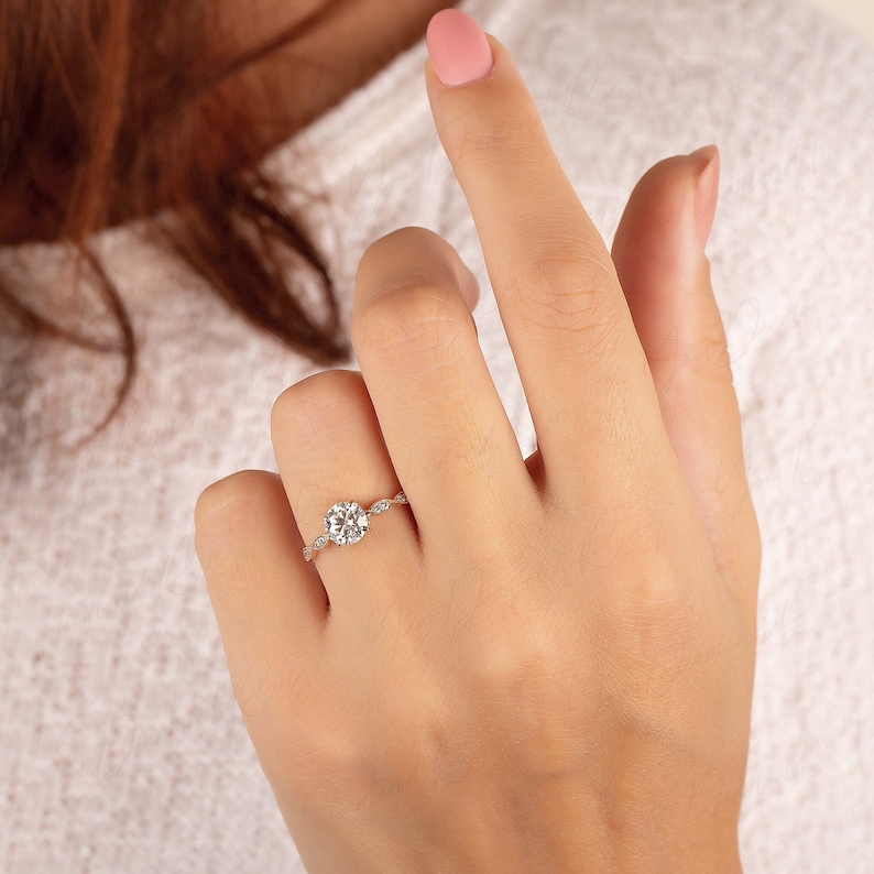 May include: A gold ring featuring a round brilliant-cut center stone held by four prongs, with a delicate band accented by small marquise-shaped stones on each side, creating a refined and symmetrical appearance on the finger.