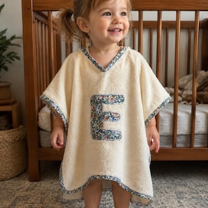 Puede incluir: Un poncho de beb&eacute; color crema con un aplique "E" con estampado floral. El poncho tiene cuello en V y est&aacute; ribeteado con tela floral azul. El ni&ntilde;o est&aacute; de pie frente a una cuna de madera.