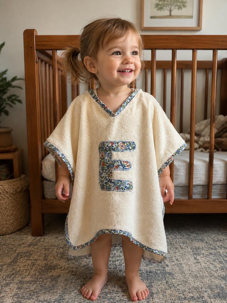 Puede incluir: Un poncho de beb&eacute; color crema con un aplique "E" con estampado floral. El poncho tiene cuello en V y est&aacute; ribeteado con tela floral azul. El ni&ntilde;o est&aacute; de pie frente a una cuna de madera.