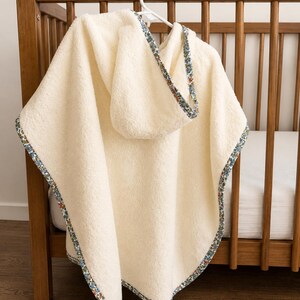 Puede incluir: Toalla de beb&eacute; con capucha de color crema con un ribete estampado floral. La toalla est&aacute; colgada de una percha blanca frente a una cuna de madera. El ribete presenta un dise&ntilde;o floral azul, rojo y verde.