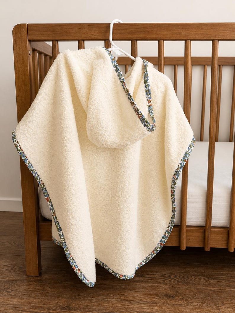 Puede incluir: Toalla de beb&eacute; con capucha de color crema con un ribete estampado floral. La toalla est&aacute; colgada de una percha blanca frente a una cuna de madera. El ribete presenta un dise&ntilde;o floral azul, rojo y verde.