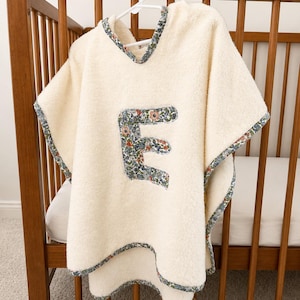 Puede incluir: Poncho de beb&eacute; color crema con una aplicaci&oacute;n de la letra "E" con estampado floral. El poncho tiene capucha y est&aacute; ribeteado con un borde de tela floral. Cuelga de una percha blanca dentro de una cuna de madera.