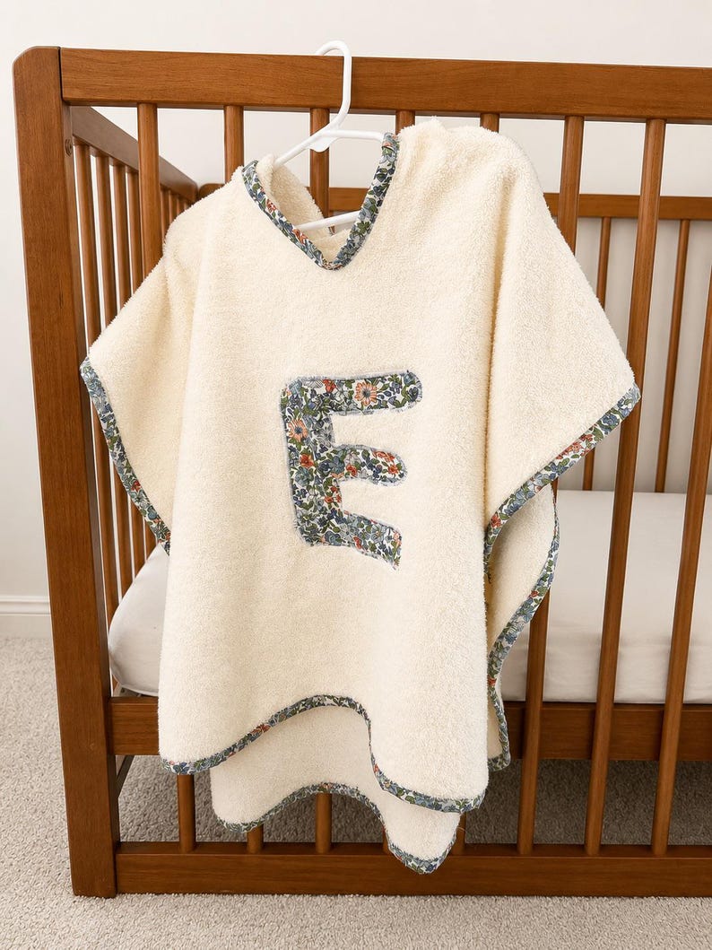 Puede incluir: Poncho de beb&eacute; color crema con una aplicaci&oacute;n de la letra "E" con estampado floral. El poncho tiene capucha y est&aacute; ribeteado con un borde de tela floral. Cuelga de una percha blanca dentro de una cuna de madera.