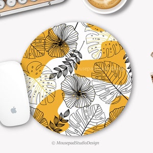 Peut inclure: Un tapis de souris rond avec un fond blanc et un motif de feuilles tropicales noires et jaunes. Le texte "Mousepad Studio Design" est en bas du tapis de souris.