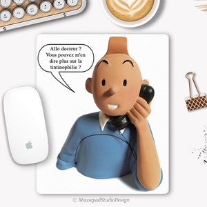 Peut inclure: Un tapis de souris avec un personnage de dessin animé au téléphone avec une bulle de dialogue qui dit "Allo docteur? Vous pouvez m'en dire plus sur la tintinophilie?" Le personnage porte une chemise bleue et a les cheveux bruns. Le tapis de souris est blanc avec une bordure noire.