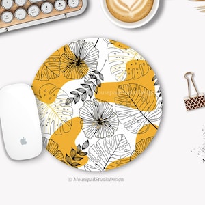 Peut inclure: Un tapis de souris rond avec un fond blanc et un motif de feuilles tropicales noires et jaunes. Le texte "Mousepad Studio Design" est en bas du tapis de souris.