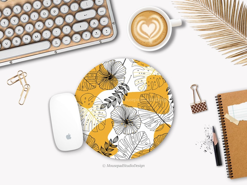 Peut inclure: Un tapis de souris rond avec un fond blanc et un motif de feuilles tropicales noires et jaunes. Le texte "Mousepad Studio Design" est en bas du tapis de souris.