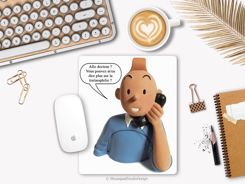 Peut inclure: Un tapis de souris avec un personnage de dessin animé au téléphone avec une bulle de dialogue qui dit "Allo docteur? Vous pouvez m'en dire plus sur la tintinophilie?" Le personnage porte une chemise bleue et a les cheveux bruns. Le tapis de souris est blanc avec une bordure noire.