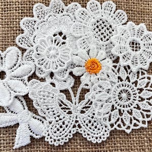 May include: White lace flower appliqués with an orange centre, arranged on a textured brown surface. The appliqués feature intricate floral designs, including daisies and other stylized flowers. Suitable for various craft projects.