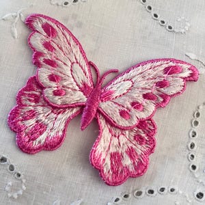 May include: Embroidered butterfly patch in pink and white. The butterfly features detailed wing patterns and a delicate, stitched appearance. The patch is likely for decorative use on clothing or accessories.