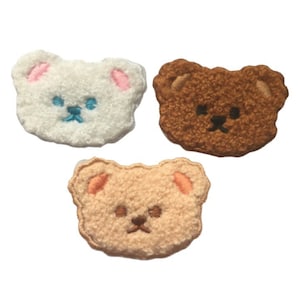 May include: Three plush bear face patches in white, brown, and tan. Each patch has embroidered facial features and pink or tan inner ears. The white bear has blue embroidered eyes and mouth.