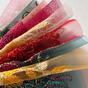 May include: A close-up of a fan of colourful lace fabric. The lace is in shades of pink, green, red, yellow, and teal. The lace has intricate embroidered designs and a mesh backing. The fabric is likely used for crafting or fashion.