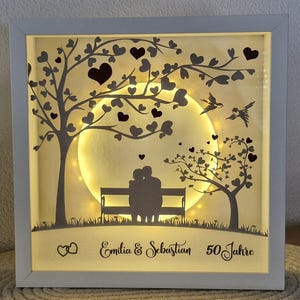 Könnte beinhalten: Eine weiße Schattenbox mit einer Silhouette eines Paares, das auf einer Bank unter einem Baum mit Vollmond und Herzen sitzt. Der Text "Emilia & Sebastian 50 Jahre" befindet sich am unteren Rand der Box.