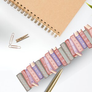 K&ouml;nnte beinhalten: Eine Sammlung dekorativer B&uuml;roklammern in Buchform in verschiedenen Farben, darunter Rosa, Lila und Grau, diagonal auf einer wei&szlig;en Oberfl&auml;che angeordnet. Ein goldener Stift, ein Spiralnotizbuch und andere B&uuml;roartikel sind ebenfalls vorhanden.