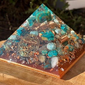 Peut inclure: Une pyramide d'orgone artisanale avec une base couleur cuivre et des accents turquoise et cuivrés. La pyramide présente des pierres incrustées, des spirales de cuivre et un motif circulaire doré. La pyramide est exposée sur une surface en bois.