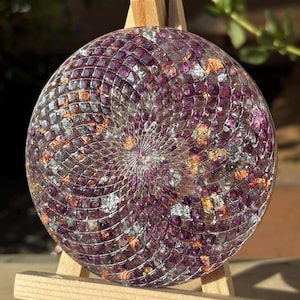 Könnte beinhalten: Ein runder, dekorativer Untersetzer mit Mosaikdesign. Der Untersetzer hat ein spiralförmiges Muster mit lila, silbernen und goldenen Akzenten. Er wird auf einer kleinen Holzstaffelei präsentiert.