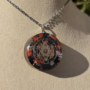 Peut inclure: Collier à chaîne argentée avec un pendentif rond. Le pendentif présente un motif géométrique doré sur un fond de pierres noires, rouges et argentées. Le collier est présenté sur une surface de ton neutre.