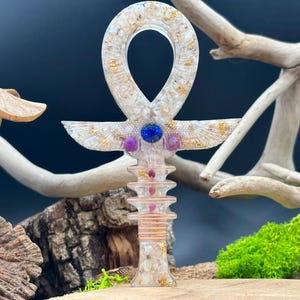 Può includere: Un ciondolo ankh in resina trasparente con scaglie d'oro e cristalli incastonati. L'ankh presenta un accento di pietre preziose blu e viola e una base a strati. Lo sfondo comprende legno e muschio verde.