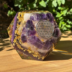 Può includere: Una piramide di orgone esagonale con cristalli di ametista, scaglie d'oro e un simbolo argentato del fiore della vita. La piramide è posta su una superficie di legno, con fogliame verde sullo sfondo. La piramide è alta circa 10 cm.