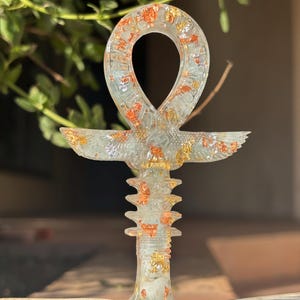 Può includere: Ciondolo Ankh in resina trasparente con inserti in foglia d'oro e rame. Il simbolo Ankh, una croce con un anello, presenta un design dettagliato con un elemento alato e una base strutturata. Il ciondolo è mostrato su uno sfondo sfocato.