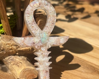 Pode incluir: Pingente ankh iridescente feito à mão com um design detalhado. O ankh apresenta um laço, asas e uma base vertical, mostrando uma mistura de cores, incluindo branco, rosa e toques de verde. O pingente está sobre um fundo de madeira natural.