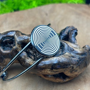 Può includere: Un braccialetto d'argento con un design circolare in bianco e nero. Il design presenta cerchi concentrici con un centro bianco.