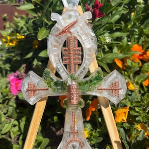 May include: A clear, cross-shaped pendant with copper wire accents and green and clear resin details. The pendant is displayed on a small wooden easel against a backdrop of colorful flowers and greenery.