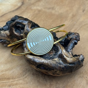 Könnte beinhalten: Ein goldenes Armreif-Armband mit einem runden, weißen und braunen Muster. Das Muster besteht aus konzentrischen Kreisen.