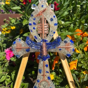 Peut inclure: Une croix Ankh en résine transparente avec des accents de lapis-lazuli bleu et des détails en fil de cuivre. L'Ankh est exposé sur un petit chevalet en bois, sur fond de fleurs colorées.