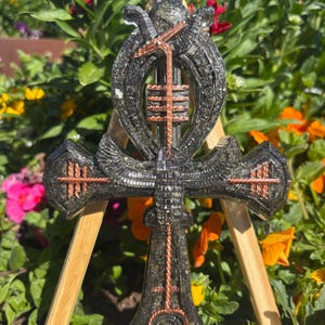 May include: A black and copper Ankh pendant with detailed design, displayed against a backdrop of vibrant flowers. The Ankh, a symbol of life, has a loop, crossbar, and vertical section, with copper wire detailing.