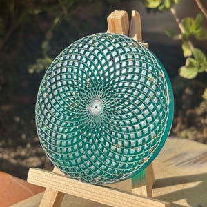 Puede incluir: Un objeto decorativo redondo y verde con un diseño geométrico en espiral. El diseño presenta una serie de cuadrados y líneas que irradian desde el centro. Hay motas doradas dispersas por todo el diseño. El objeto se muestra en un pequeño caballete de madera.